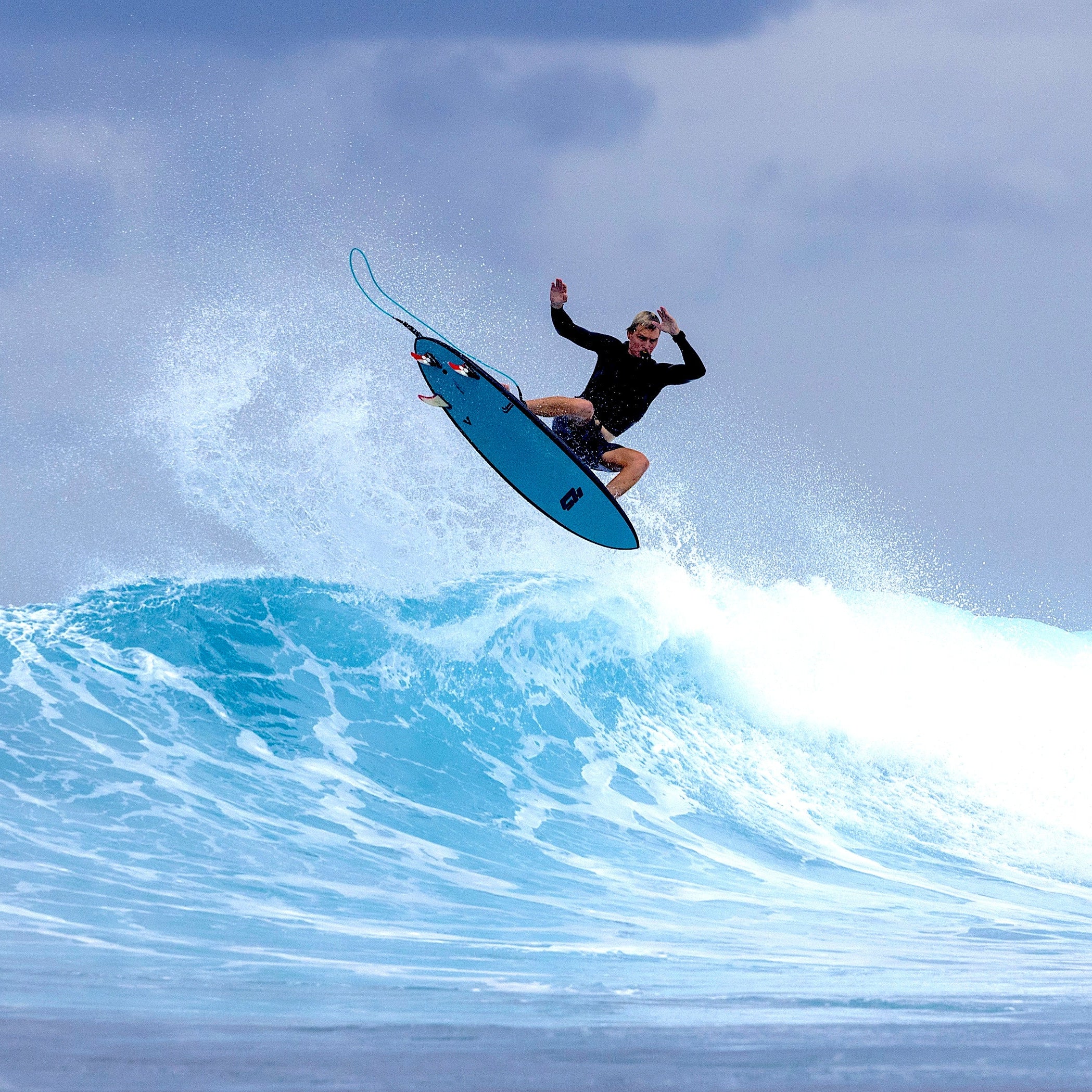 Surfer performing a trick on a blue surfboard over a wave with a cloudy sky.